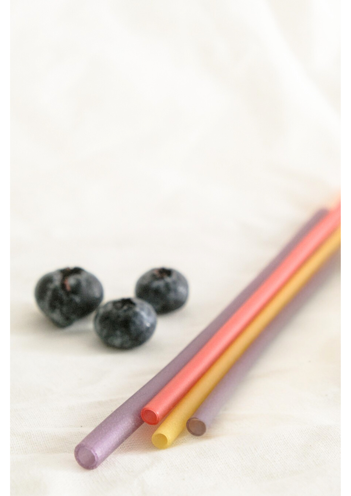 Three colorful rice straws in purple, yellow, and pink, placed next to three blueberries on a light fabric background.