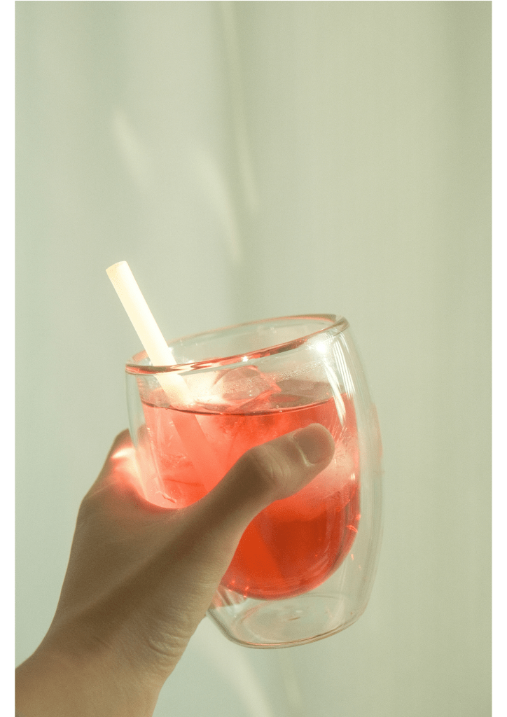 A hand holding a transparent glass cup filled with a pink drink and ice, with a rice straw inside.